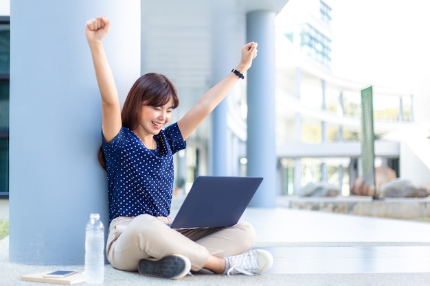 Jovem mulher de negócios asiática levanta os braços com entusiasmo enquanto olha para o seu computador portátil