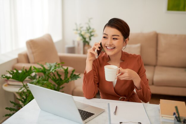 Jovem mulher de negócios asiática segurando uma caneca e usando o smartphone enquanto está sentada na mesa do escritório com o laptop