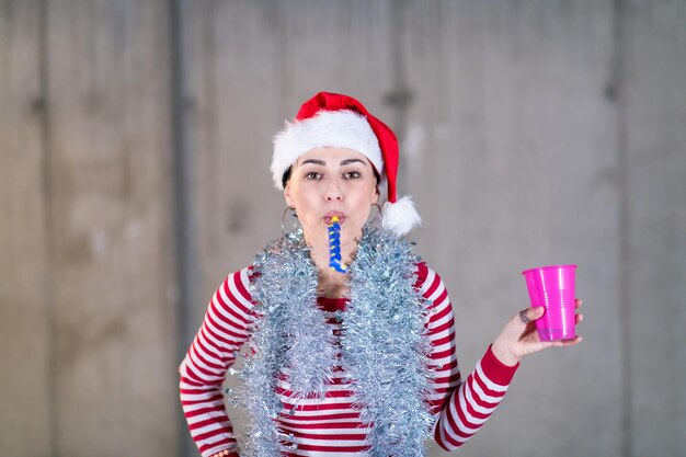 jovem mulher de negócios casual feliz usando um chapéu vermelho e soprando apito de festa enquanto dançava durante a festa de ano novo em frente a parede de concreto