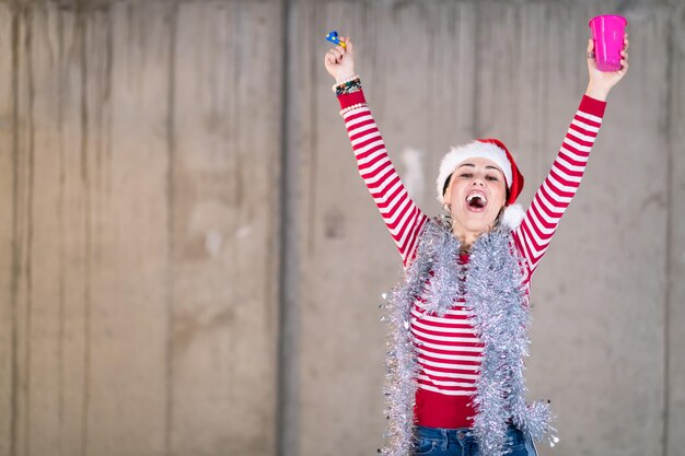 jovem mulher de negócios casual feliz usando um chapéu vermelho e soprando apito de festa enquanto dançava durante a festa de ano novo em frente a parede de concreto