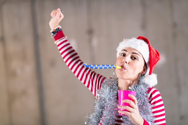 jovem mulher de negócios casual feliz usando um chapéu vermelho e soprando apito de festa enquanto dançava durante a festa de ano novo em frente a parede de concreto