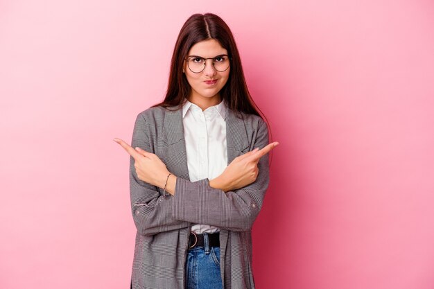 Jovem mulher de negócios caucasiana isolada na parede rosa aponta para o lado, está tentando escolher entre duas opções.