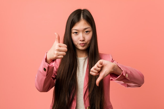 Jovem mulher de negócios chinesa vestindo um terno rosa mostrando os polegares para cima e para baixo, difícil escolher o conceito