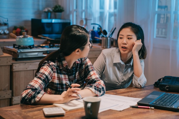 jovem mulher de negócios feminino de volta para casa tarde da noite sentado na mesa da cozinha em casa enquanto companheiro de quarto usando o computador portátil para calcular as contas. duas senhoras asiáticas fofocam conversando falando à meia-noite.