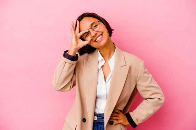 Jovem mulher de negócios isolada na parede rosa animada mantendo um gesto de ok no olho