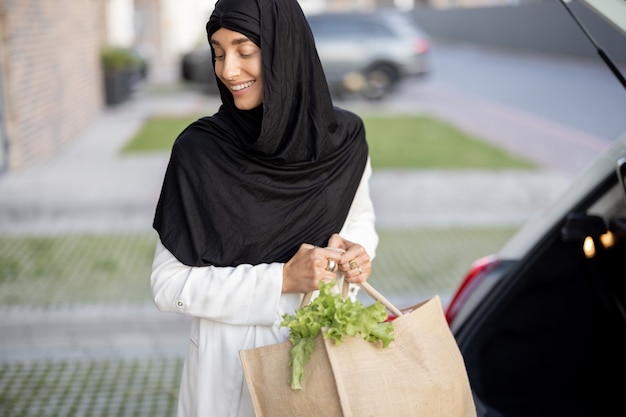 Jovem mulher de negócios muçulmana em um hijab indo para casa com mantimentos, em pé com uma sacola de compras na varanda de sua casa