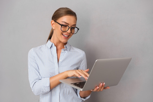 Jovem mulher de negócios posando isolado sobre o fundo da parede cinza, usando o computador portátil.