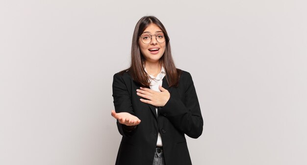 Jovem mulher de negócios se sentindo feliz e apaixonada, sorrindo com uma mão ao lado do coração e a outra esticada na frente