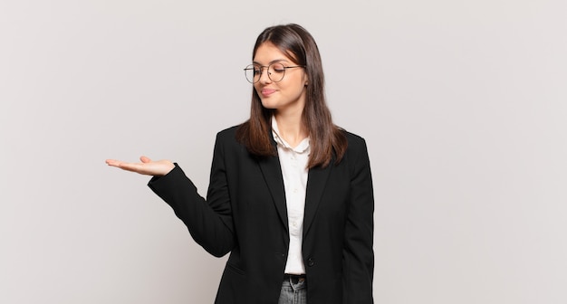 Jovem mulher de negócios se sentindo feliz e sorrindo casualmente, olhando para um objeto ou conceito seguro ao lado