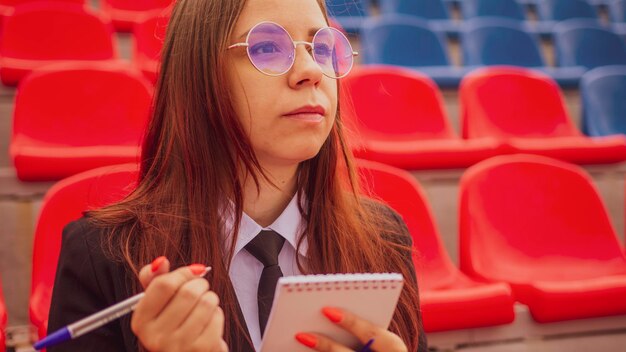 Jovem mulher de óculos com caneta de bloco de notas sentada nas arquibancadas do estádio sozinha Jornalista feminina escrevendo notas durante as competições no estádio de rua