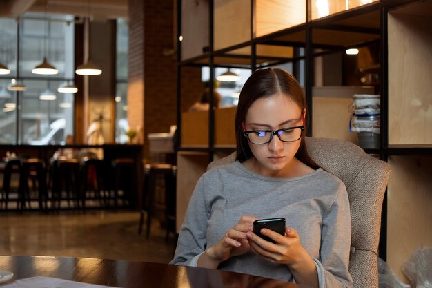 Jovem mulher de óculos sentada à mesa e navegando nas mídias sociais no smartphone enquanto descansa