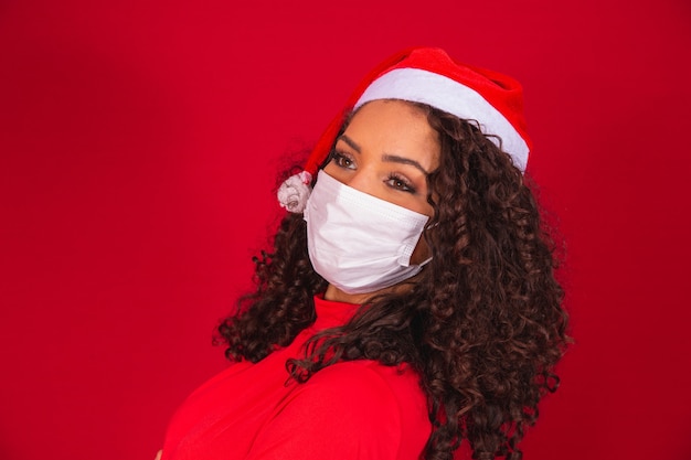 Jovem mulher de Papai Noel com chapéu de Natal usando o vírus covid-19 coronavirus salvo máscara isolada no estúdio de fundo vermelho. Conceito de feriado de celebração de ano novo feliz.