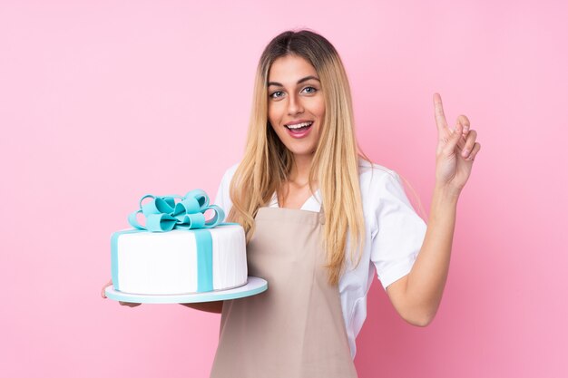 Jovem mulher de pastelaria uruguaia com um bolo grande sobre parede rosa isolada apontando uma ótima idéia