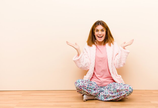 Jovem mulher de pijama, sentada em casa, sentindo-se feliz, animada, surpresa ou chocada, sorrindo e espantada com algo inacreditável