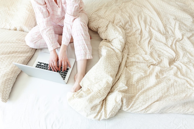 Jovem mulher de pijama, sentada na cama em casa trabalhando usando no laptop