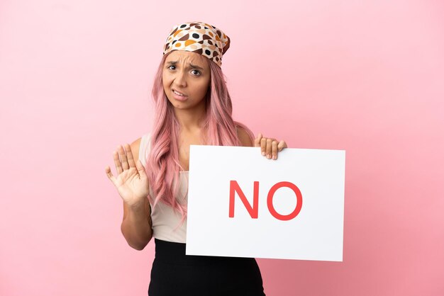 Jovem mulher de raça mista com cabelo rosa isolada em um fundo rosa segurando um cartaz com o texto NÃO e fazendo o sinal de pare