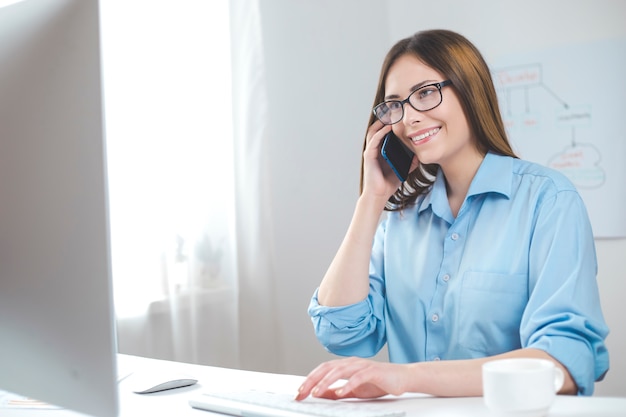 Jovem mulher de sorriso que fala no telefone ao sentar-se no computador no local de trabalho no escritório.