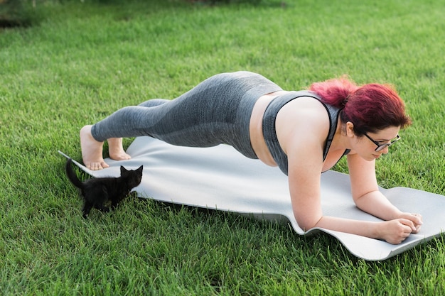 Jovem mulher de tamanho grande em top esportivo e leggings em pé na prancha no tapete de ioga passando tempo em