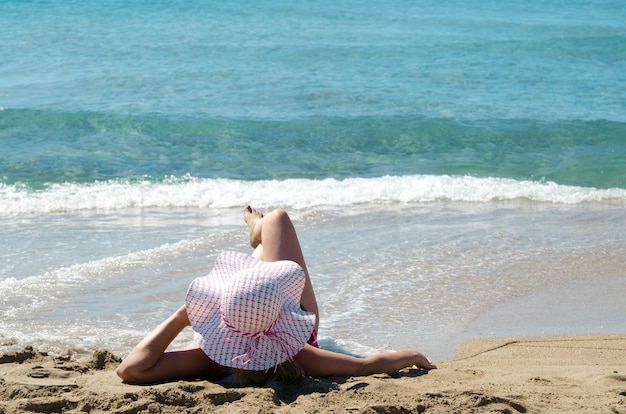 Jovem mulher de volta, deitada com chapéu de sol na praia de areia e aproveite as férias de verão. Bronzeamento no belo dia.