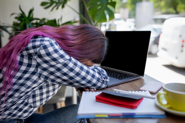 Jovem mulher deitada de braços sobre a mesa no café na frente do laptop com uma xícara de café, com sono. Foto traseira de uma aluna aprendendo on-line via laptop antes de suas palestras.