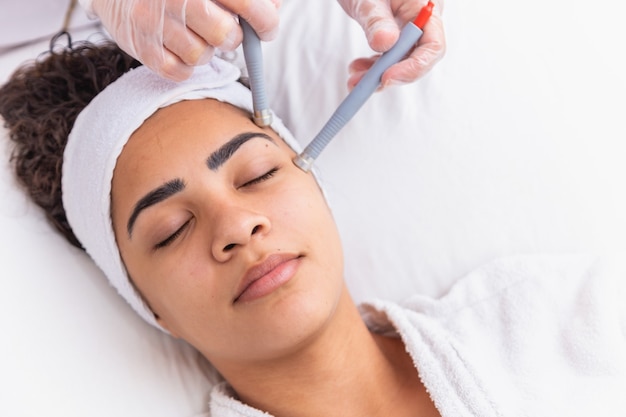 Jovem mulher deitada na mesa do cosmetologista durante o procedimento de rejuvenescimento. O cosmetologista cuida da juventude e do bem-estar da pele do pescoço e do rosto. Procedimento de limpeza da face de hardware.