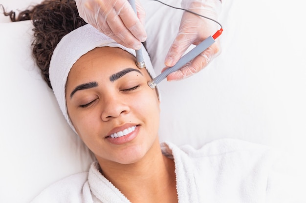 Jovem mulher deitada na mesa do cosmetologista durante o procedimento de rejuvenescimento. o cosmetologista cuida da juventude e do bem-estar da pele do pescoço e do rosto. procedimento de limpeza da face de hardware.