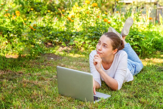 Jovem mulher deitada no gramado da grama verde no parque da cidade, trabalhando no computador pc portátil.