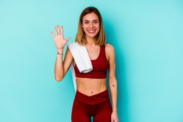 Jovem mulher desportiva, caucasiana, isolada na parede azul, sorrindo alegre mostrando o número cinco com os dedos