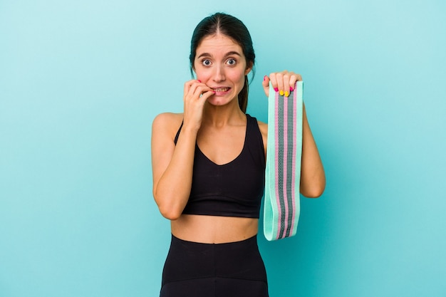 Jovem mulher desportiva, caucasiana, segurando elásticos isolados em fundo azul, roendo as unhas, nervosa e muito ansiosa.