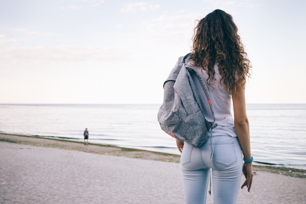 Foto jovem mulher desportiva com cabelos cacheados, vestindo jeans e uma mochila em pé na praia e olhando para o mar