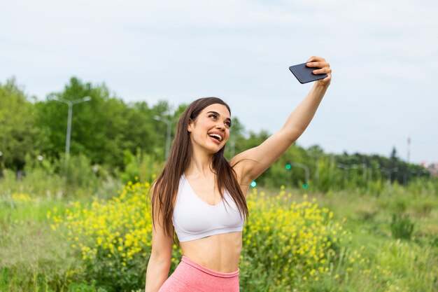 Jovem mulher desportiva tomando uma selfie no parque. Ela está olhando para a câmera.