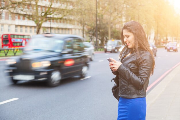 Jovem, mulher, digitando em seu telefone inteligente por uma estrada movimentada em londres