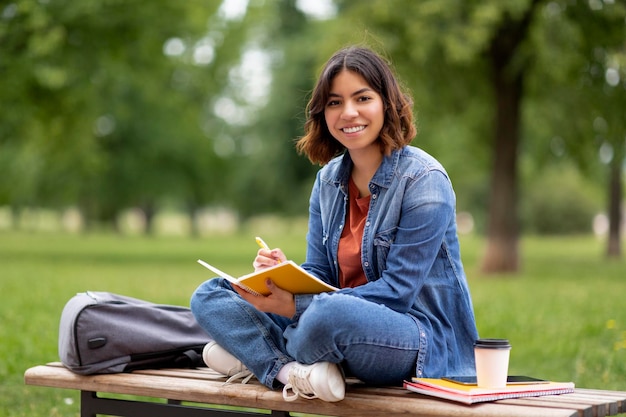 Jovem mulher do Oriente Médio escrevendo no caderno enquanto está sentada no banco ao ar livre