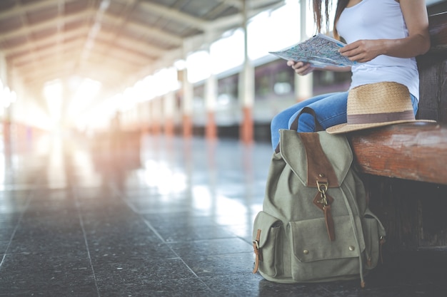 Jovem mulher dos viajantes com a trouxa que olha guarda um mapa na estação de trem. Dia do turismo.