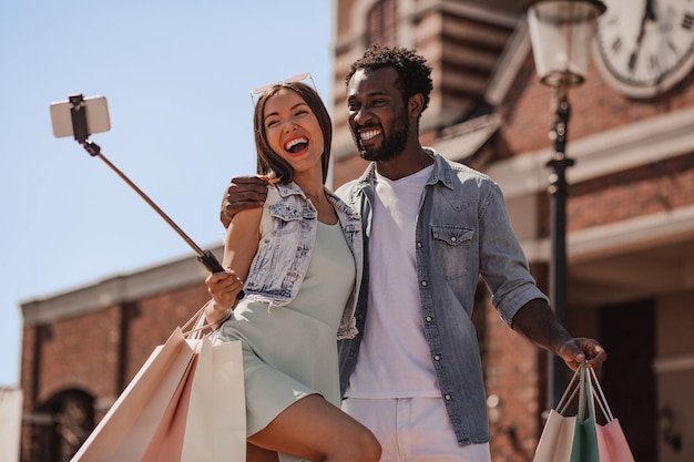 Jovem mulher e homem com sacolas de compras e tomando selfie com smartphone usando selfiestick na rua