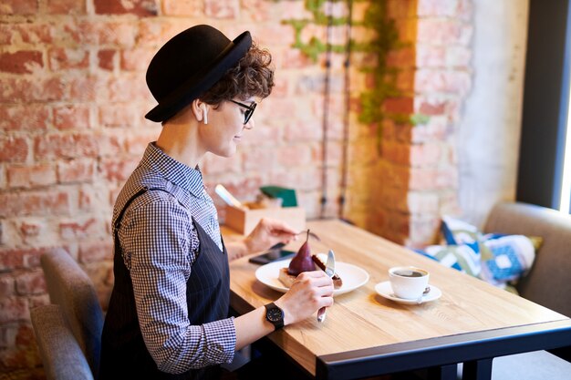 Jovem mulher elegante com airpods sentado no café no intervalo do almoço e comendo sobremesa deliciosa por tabela