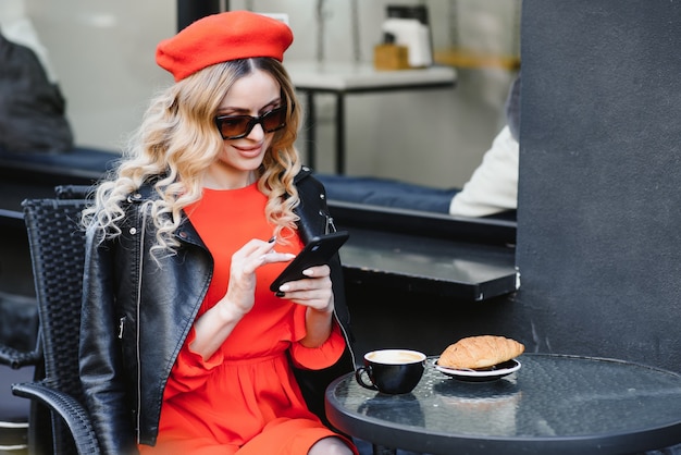 Jovem mulher elegante com boina vermelha, tomando um café da manhã francês com café e croissant, sentada ao ar livre no terraço do café