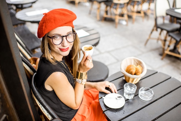 Jovem mulher elegante com boina vermelha, tomando um café da manhã francês com café e croissant, sentada ao ar livre no terraço do café