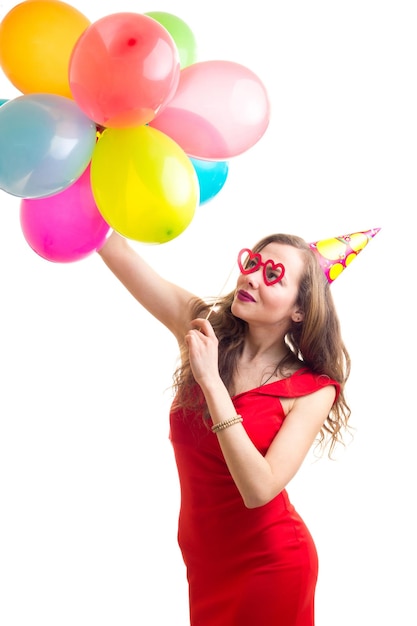 Jovem mulher elegante com longos cabelos castanhos vestido vermelho com chapéu comemorando segurando muitos balões coloridos e cartão de óculos em fundo branco no estúdio.