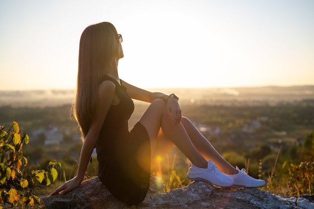 Jovem mulher elegante em um vestido preto curto, sentado em uma rocha relaxante ao ar livre em noite de verão. Mulher elegante, apreciando o pôr do sol quente na natureza.