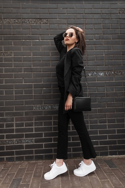 Jovem mulher elegante em uma jaqueta preta longa na moda em jeans vintage em uma camiseta preta da moda com bolsa de couro preta elegante perto da parede de tijolo ao ar livre. Moda feminina moderna. Primavera Verão.