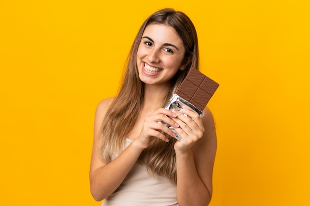 Jovem mulher em amarelo isolado, tomando um comprimido de chocolate e feliz