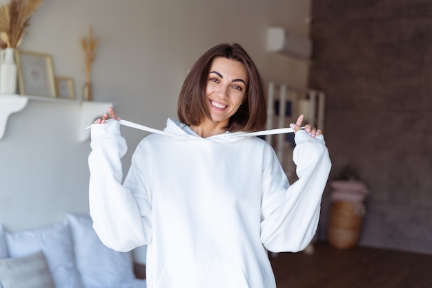 Jovem mulher em casa no quarto com um moletom branco quente, feliz, posando, sorridente, aconchegante noite de inverno