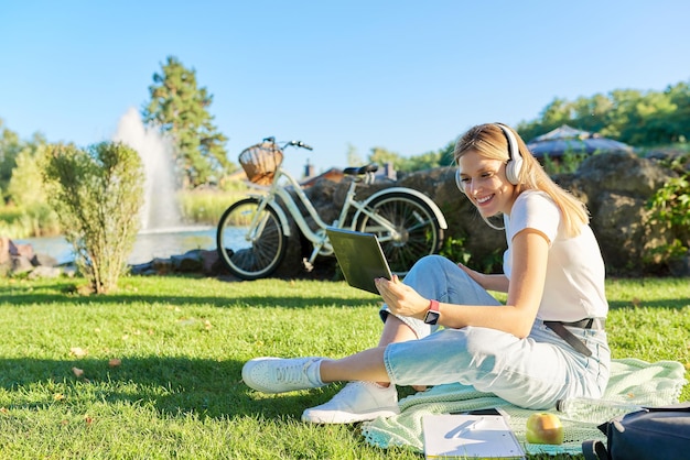 Jovem mulher em fones de ouvido com tablet digital olhando na tela do vídeo do gadget chamada Mulher sentada no gramado verde no parque no dia ensolarado de verão