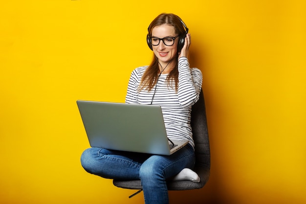 Jovem mulher em fones de ouvido, ouvindo música e sentado na cadeira e segurando o laptop sobre fundo amarelo isolado.