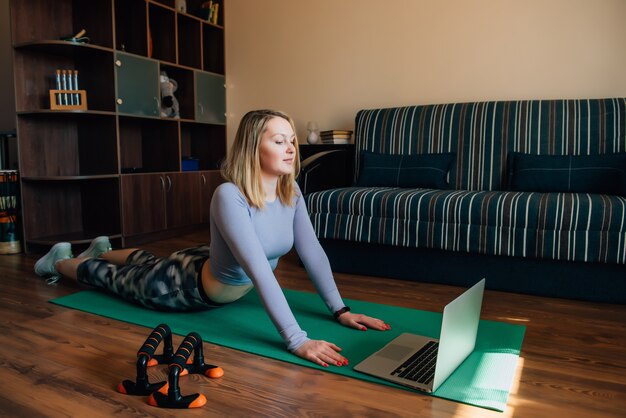 Jovem mulher em pose de ioga assistindo aula on-line em casa durante a quarentena. Garota magro atraente no sportswear fazendo exercícios de fitness na esteira.