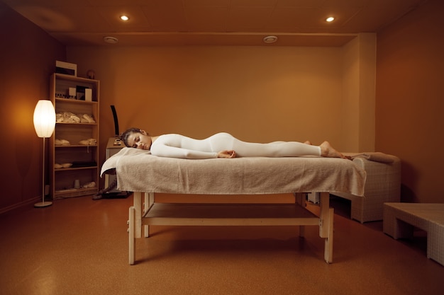 Jovem mulher em roupa de massagem relaxante na mesa. Massagem e relaxamento, cuidados com o corpo e a pele. Mulher deitada em salão de spa