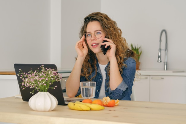 Jovem mulher em roupas casuais, sentada à mesa com notebook e frutas na mesa enquanto fala no celular na cozinha