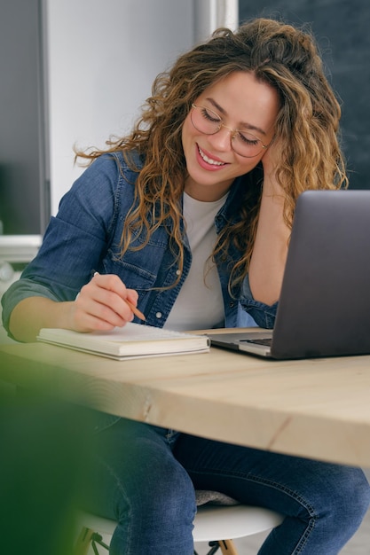 Jovem mulher em roupas casuais, sentada à mesa com o laptop e trabalhando no projeto enquanto faz anotações no planejador