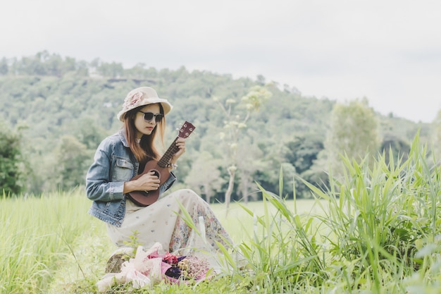 Jovem mulher em um campo, óculos de sol arredondados e chapéu de palha e ukulele se divertindo ao ar livre.